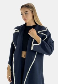 Marineblauer Mantel mit weißer dekorativer Naht, mit weitem Kragen und übergroßen Ärmeln. Strukturierter Stoff, offene Vorderseite und lockere Passform.