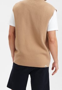 Beige ärmelloser Strickpullover mit strukturierter Oberfläche, über einem weißen T-Shirt getragen. Zusätzliches Detail ist ein kleines Etikett am Saum.