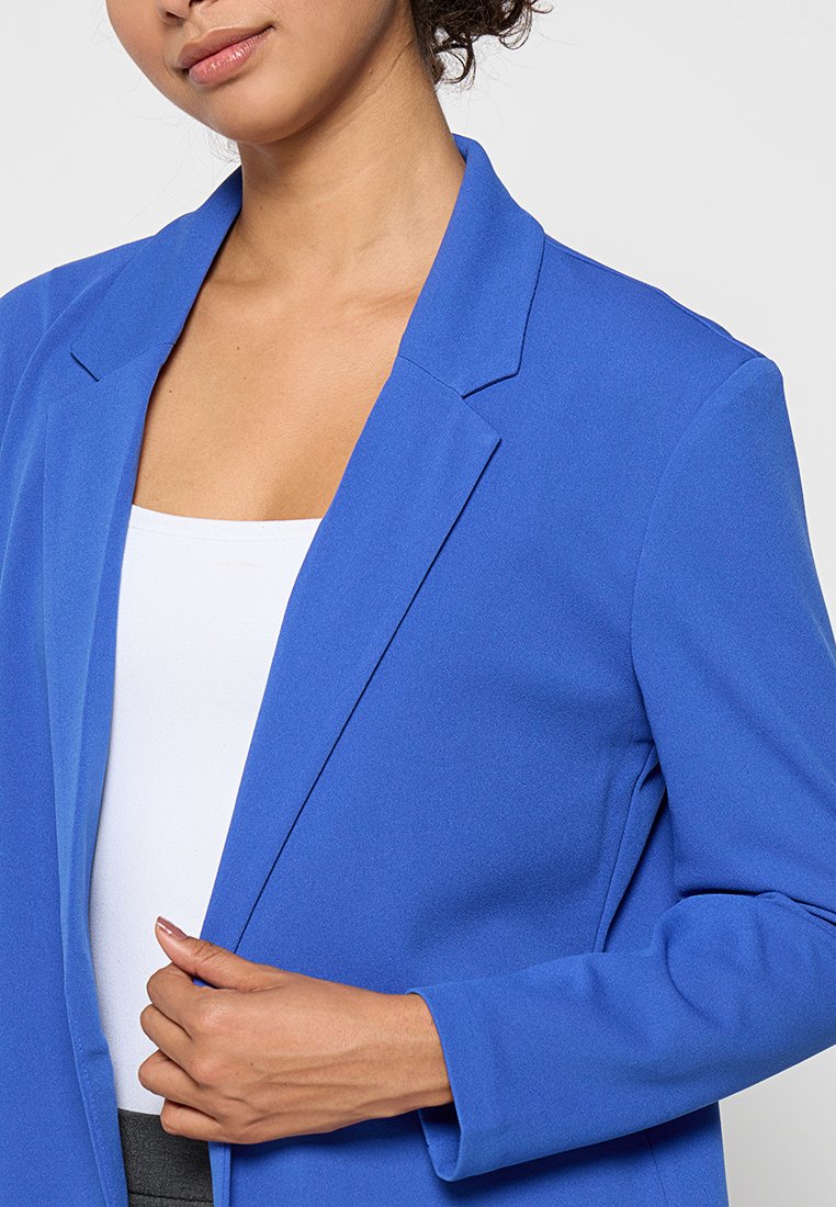 Blauer Blazer aus Stretchstoff mit einem Ein-Knopf-Design, Reverskragen und taillierter Passform. Über einem weißen Oberteil getragen.