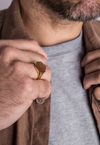 Gold-toned hexagonal ring with wood inlay, featuring a smooth surface and triangular accents, worn on a hand gripping a brown corduroy jacket.