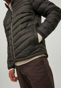 Svart puffjakke med quiltet design, ribbstrikkede mansjetter og glidelås. Brukt over en lys genser og brune bukser.