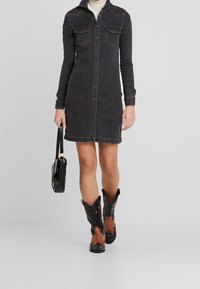 Mekko mustasta denimestä nappikiinnityksellä, pitkät hihat, rintataskut ja hieman kaareva helma, yhdistettynä mustiin cowboy-kengiä ja mustaan käsilaukkuun.