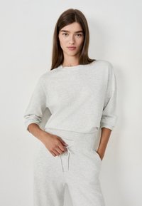 Young woman with long brown hair wearing light grey sweatshirt and matching sweatpants, posing against a plain white background.