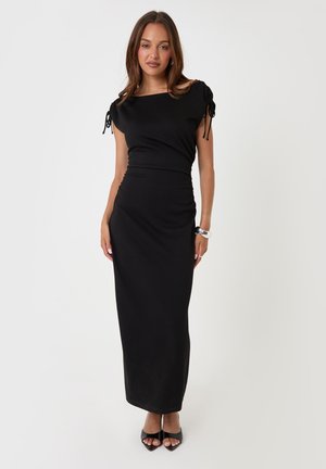 Femme portant une longue robe noire à manches froncées, debout les mains détendues, chaussée de talons ouverts noirs et portant une montre au poignet gauche.