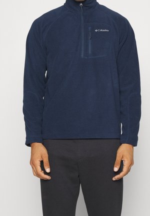 Man die een navyblauwe Columbia fleece trui met ritszak op de borst draagt, staand tegen een effen lichte achtergrond.