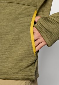 Olivovo zelený dlhý rukáv top s textúrovanou látkou, ktorý má žltý akcent pri vrecku. Hladké rukávy a voľný strih.