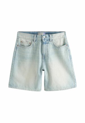 Person løfter hvid top for at vise lyseblå denimshorts med frontlommer og knaplukning mod en ensfarvet baggrund.