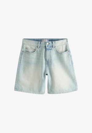 Person løfter hvid top for at vise lyseblå denimshorts med frontlommer og knaplukning mod en ensfarvet baggrund.
