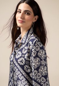 Blusa blu navy con un motivo floreale bianco. Presenta un colletto e una vestibilità morbida, realizzata in un tessuto liscio con una leggera lucentezza.