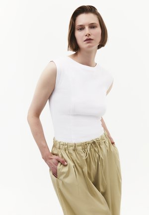 Frau trägt ein ärmelloses weißes Oberteil und lockere beige Hosen, die Hände in den Taschen, steht vor einem schlichten weißen Hintergrund.