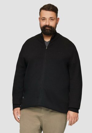 Zwarte ritscardigan in textiele stof, met een gebreide kraag en manchetten. Wordt gedragen met een grijze T-shirt eronder en neutrale broek.