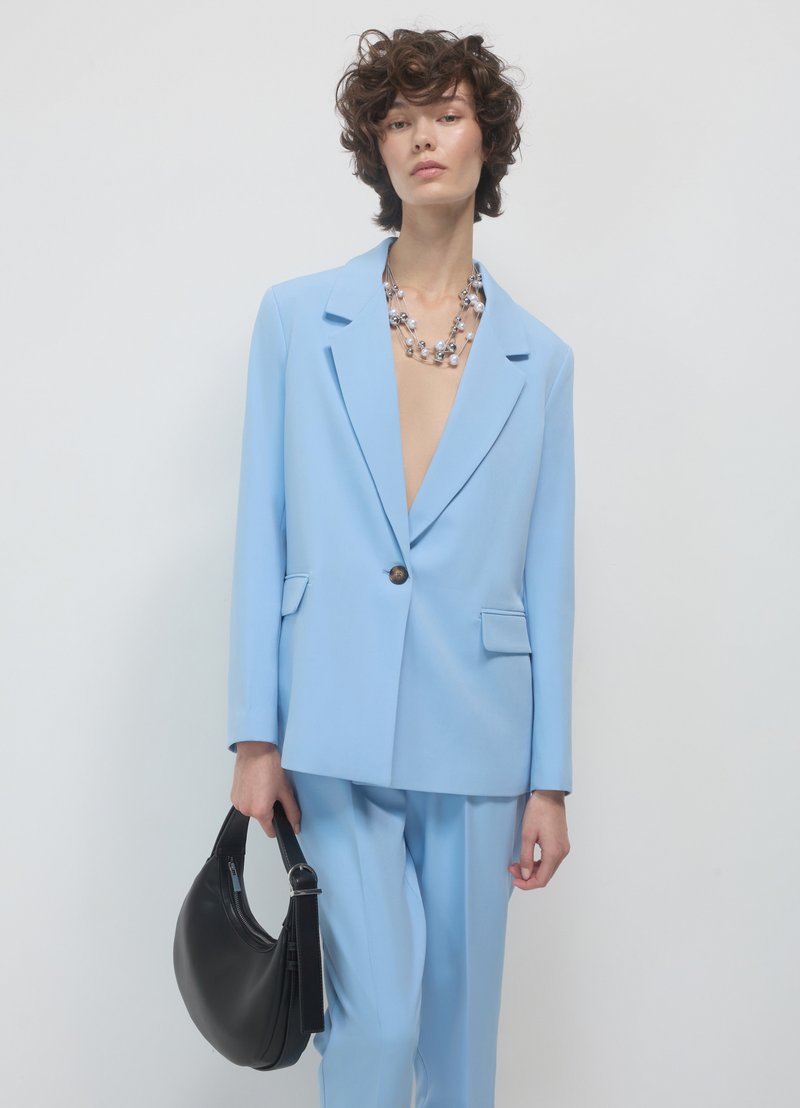 Blazer bleu clair et pantalon assorti, avec un seul bouton, des poches à rabat, accompagné d'un sac noir et d'un collier multicolore.