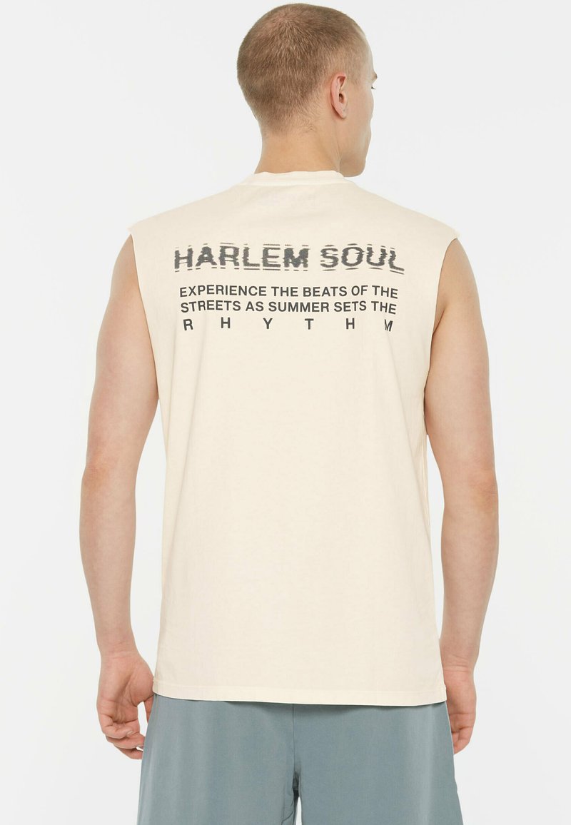 Beige sleeveless t-shirt with text design on the back, including "HARLEM SOUL" and other phrases in black. Soft cotton fabric.