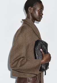 Woman in brown wool coat and leather pants holding a black woven clutch bag against a white background.