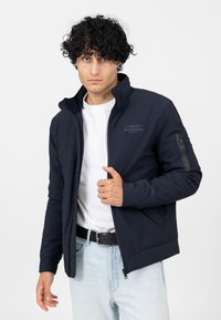 Navy Softshelljacke mit hohem Kragen, seitlichen Reißverschlusstaschen und dezentem Logo auf der Brust. Mit elastischen Bündchen und figurnaher Passform.