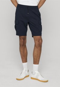 Shorts cargo bleu marine avec deux poches latérales, ceinture élastique et fermeture à cordon, associés à des baskets blanches et des chaussettes côtelées.