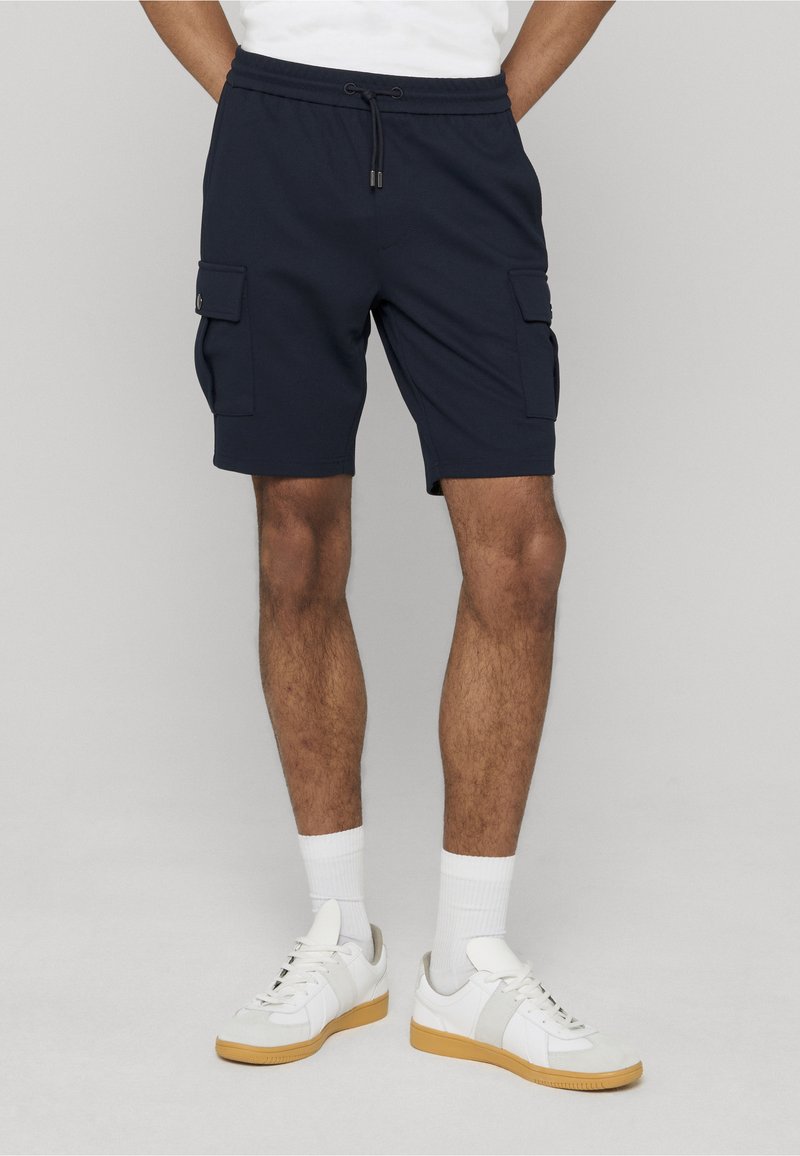 Shorts cargo bleu marine avec deux poches latérales, ceinture élastique et fermeture à cordon, associés à des baskets blanches et des chaussettes côtelées.