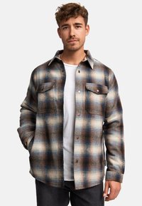 Camicia di flanella a quadri marrone e blu con due tasche sul petto, davanti abbottonata e con una texture morbida. Indossata sopra una maglietta bianca e jeans grigi.