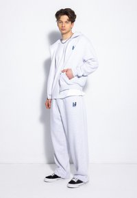 Jasnoszara bluza z kapturem, z kieszenią z przodu i logo; zestawiona z dopasowanymi spodniami dresowymi z logo po boku i czarnymi sneakersami.