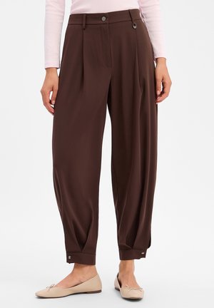 Vrouw draagt bruine losse enkellange broek met knoopmanchetten en beige ballerina's tegen een effen achtergrond.