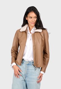 Vrouw in een tan leren jasje met witte bontkraag, witte blouse en lichtblauwe spijkerbroek, staande tegen een effen achtergrond.