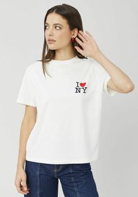 T-shirt en coton blanc avec un graphisme "I ❤️ NY" en rouge et marine, manches courtes, coupe décontractée. Porté avec un jean bleu foncé.