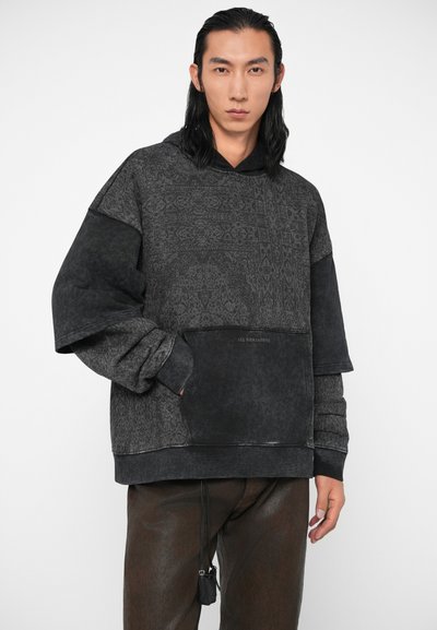 Mlađi muškarac s dugom tamnom kosom nosi široku tamnu kapu s uzorkom i tamne hlače, stoji s jednom rukom u džepu dukserice.