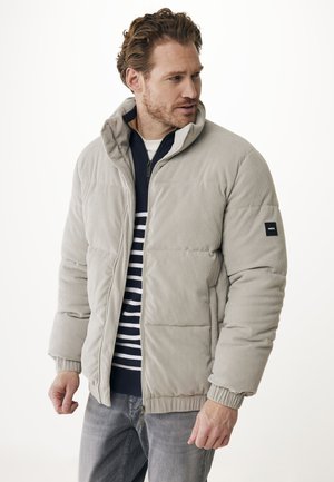 Uomo che indossa una giacca imbottita in corduroy grigio chiaro sopra un maglione a righe blu e bianche, che guarda a destra, in piedi contro uno sfondo neutro.
