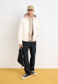 Light cream jacket over a beige hoodie, paired with dark cargo pants and black sneakers. A green cap and a small black bag complete the look.
