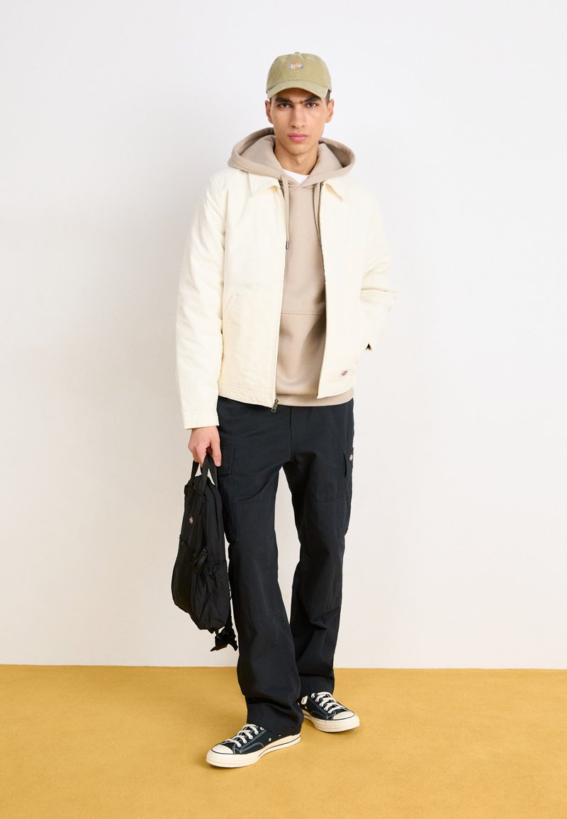 Veste crème claire sur un sweat à capuche beige, associée à un pantalon cargo noir et des baskets noires. Une casquette verte et un petit sac noir complètent le look.