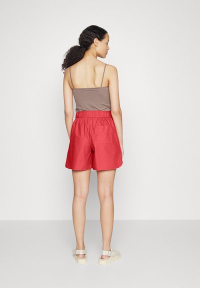 Røde shorts med elastisk talje, med en enkelt baklomme. Kombinert med en brun tanktop og beige sandaler. Minimalistisk design.