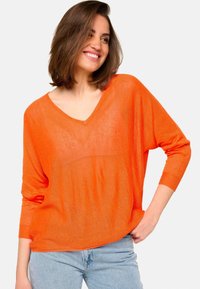 Femme souriante avec des cheveux bruns jusqu'aux épaules portant un pull orange à col en V et un jean bleu clair, la main reposant sur la hanche.