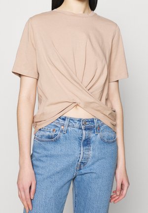 Vaaleabeige puuvillainen t-paita, jossa on solmittu solmu vyötäröllä, yhdistetty korkeavyötäröisiin vaaleansinisiin farkkuihin. Lyhyet hihat, pyöreä kaula-aukko.