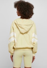 Gul huva-jacka med vita ränder på ärmarna och en avslappnad passform. Materialet verkar lätt; kombinerad med matchande ljusgula shorts.