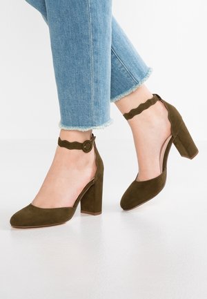 Füße in olivgrünen Wildleder-Pumps mit Blockabsatz und wellenförmigen Knöchelriemen, kombiniert mit hellblauen Jeans mit ausgefransten Säumen.