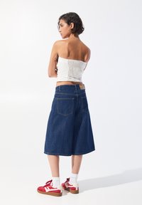 Denim-Culottes, dunkelblau, mit einer mittelhohen Taille und Gesäßtaschen. Kombiniert mit einem weißen Smoking-Oberteil und roten Sportschuhen mit weißen Akzenten.