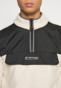 Fleecejacka med hög krage, i svart och krämfärgat, med frontdragkedja och en logotypapplikation på bröstet.