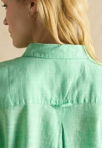 Groen geruit shirt met een kraag, plooidetail aan de achterkant en textuurstof. Gouden oorbellen zijn zichtbaar op de voorgrond.