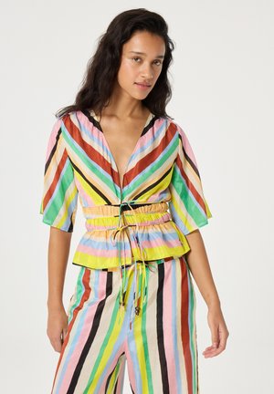 Woman wearing colorful vertical and diagonal striped blouse with drawstring waist and matching pants, standing against plain background.
