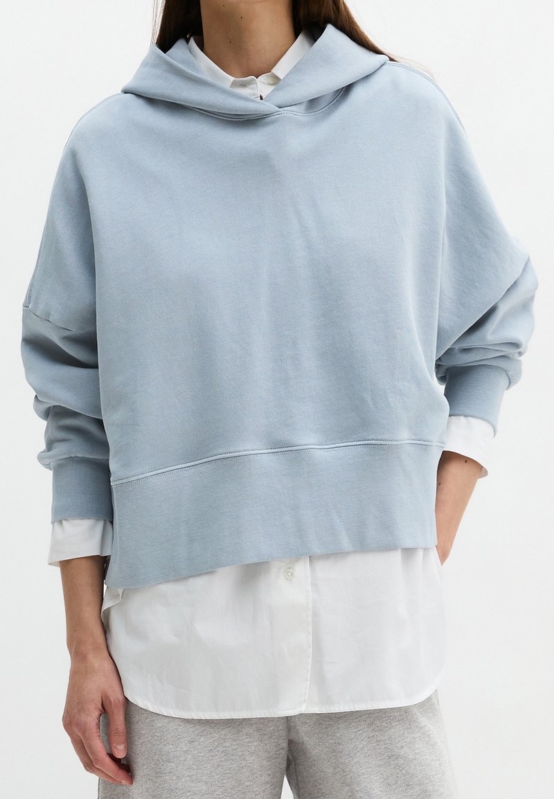 Personne portant un sweat à capuche court bleu clair sur une chemise blanche à boutons et un pantalon de survêtement gris, debout avec une main dans la poche.