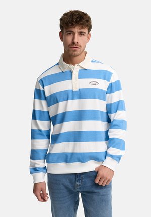 Mężczyzna ubrany w koszulkę rugby w poziome niebiesko-białe paski z białym kołnierzem i niebieskie dżinsy, stojący na tle jednolitego tła.