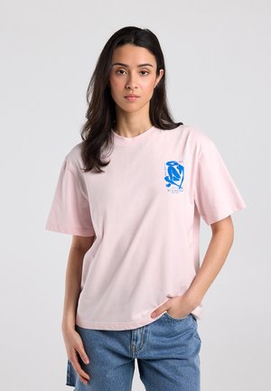 Woman with long dark hair wearing a light pink T-shirt with a small blue graphic on the chest and blue jeans, standing against a plain background.