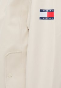 Tejido beige con un bolsillo cosido y un botón, que presenta un logotipo rectangular de Tommy Jeans en azul marino, blanco y rojo en la parte superior derecha.