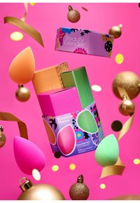 Il Beauty Blender Essentials Set include spugne nei colori rosa, verde e arancione, confezionate in una scatola colorata con motivi decorativi. Lo sfondo è rosa.