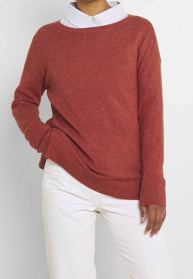 Personne portant un pull en maille couleur rouille sur une chemise blanche à col, associé à un pantalon blanc, posant avec une main dans la poche.