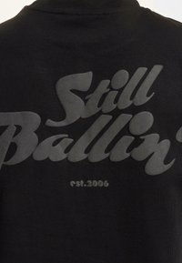 Černé tričko s výrazným lesklým nápisem "Still Ballin'" ve stylizovaném písmu, s "est. 2006" pod ním, ukazující hladkou texturu látky.