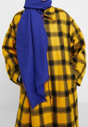 Personne portant un manteau à carreaux jaunes et noirs, les mains dans les poches, avec une épaisse écharpe bleue enroulée autour du cou.