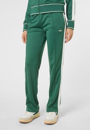 Person iført grønne træningsbukser med hvide sidelister, hvide sneakers og matchende grøn jakke med hvide detaljer.