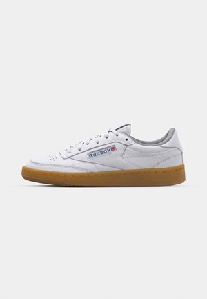 Balti ādas sporta apavi ar pelēku mīkstinošu apmalīti, auklu slēgšanas sistēmu un gumijas zoli. Sānos ir zils un sarkans Reebok logo.