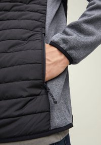 Black quilted vest with a gray knit section. Features zippered pockets and a drawstring at the hem. Smooth texture throughout.